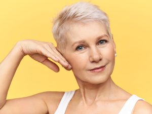portrait of attractive middle aged caucasian woman in tank top touching cheek applying anti wrinkle moisturizing cream looking at camera with smile